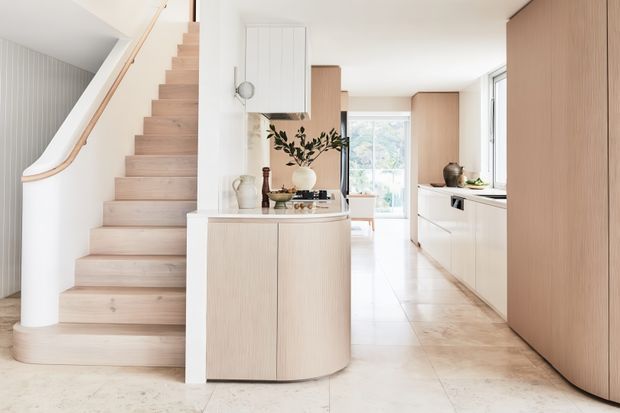 Bright open-plan kitchen and entry interior with a pale timber staircase, curved timber handrail, light wood cabinetry, white benchtops, and a freestanding curved island or console. The kitchen extends along the right side, while a hallway and glazing provide depth toward the rear. The space has a clean, restrained, contemporary feel with soft natural light and pale neutral finishes.