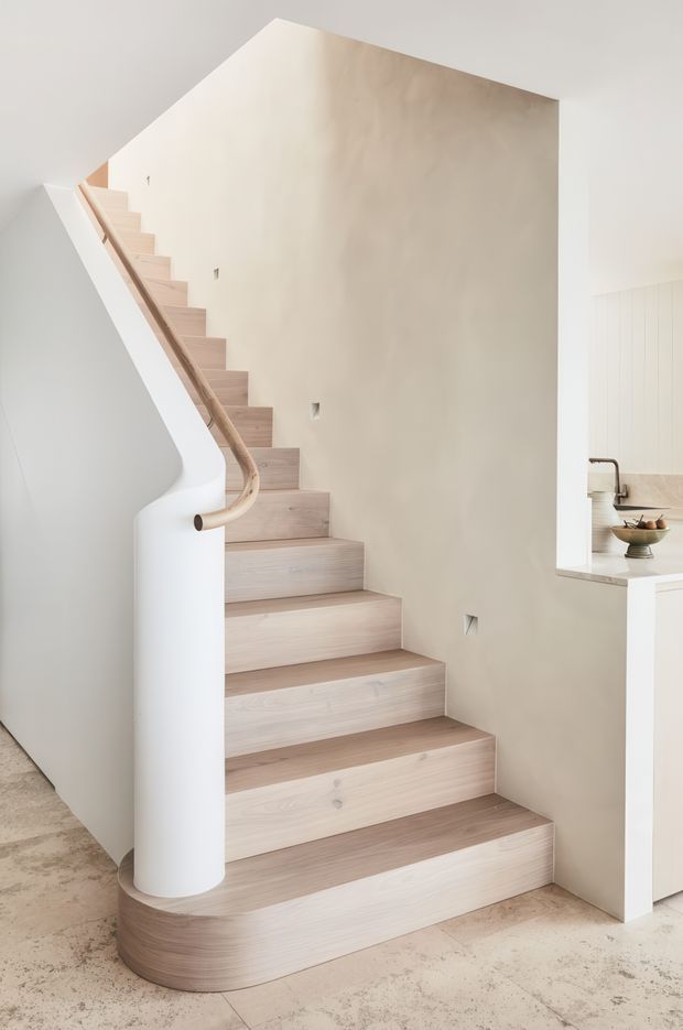 A bright minimalist interior focused on a sculptural staircase. Pale timber stair treads and a curved timber handrail rise alongside a smooth white curved balustrade and rounded newel-like column. The surrounding walls are warm off-white, with a pale concrete or stone-look floor. A portion of a light kitchen with white cabinetry and a pale benchtop is visible at right.