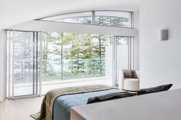 Bright contemporary bedroom with a low-profile bed facing expansive floor-to-ceiling glazing. The room features a curved clerestory-style window band, slim pale window frames, white walls, light timber flooring, and a neutral upholstered armchair. Trees and a glass balustrade are visible outside, creating an airy connection to the landscape.