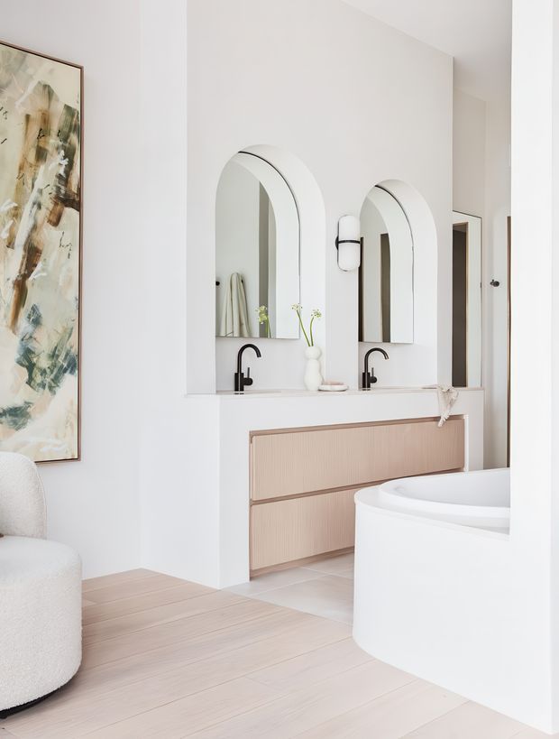 Bright, refined bathroom with a double vanity featuring pale timber fluted drawers, white benchtop, black curved tapware, and twin arched mirrors. A freestanding white bathtub occupies the foreground, while pale timber-look flooring, white walls, recessed architectural niches, and soft neutral furnishings create a clean contemporary interior. An adjacent upholstered seating area and artwork are visible at the left edge.