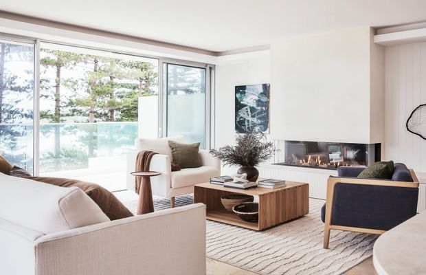 Bright, refined living room with large corner glazing, a built-in fireplace and television wall, pale upholstered seating, a sculptural timber coffee table, and a textured neutral rug. The space opens to a balcony or terrace with views of trees and water beyond.