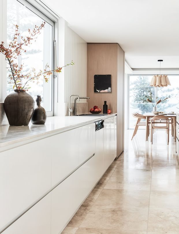 Bright, refined contemporary kitchen viewed toward an adjoining dining area. The kitchen features a long run of sleek white handleless cabinetry, a pale stone benchtop, integrated sink and tap, and a warm vertical timber-panelled feature wall with a small dark artwork. Large windows bring in abundant natural light and overlook trees. A light timber dining table and chairs sit beyond the kitchen, beneath a sculptural pendant light. The floor is finished in large pale beige stone-look tiles, while ceramic vessels and a branch arrangement add organic character.