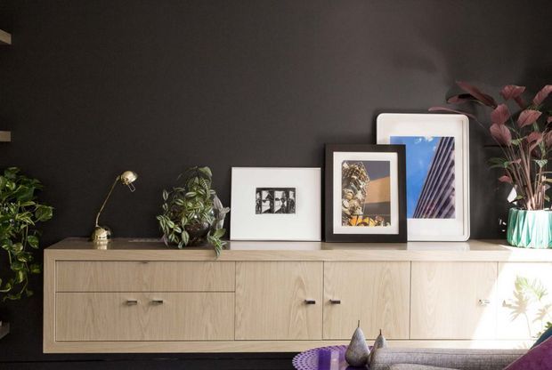 A styled interior vignette featuring a long light-oak built-in or freestanding sideboard against a dark charcoal wall. The cabinet has simple flat-front drawers and doors with small dark handles, while framed artwork, potted plants, and a brass-toned adjustable lamp sit on top. The lower foreground includes part of a sofa and decorative objects. The composition is moody, refined, and contemporary, with strong contrast between the dark wall and pale timber joinery.