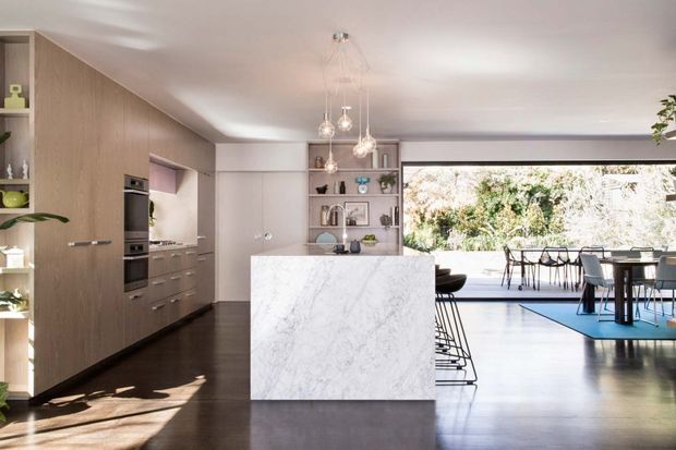 Open-plan contemporary kitchen and dining area with warm timber cabinetry, dark timber flooring, a large white marble-look island, pendant lighting, and extensive glazing opening to a garden or outdoor setting. A dining table and chairs are visible beyond the island, with integrated shelving and appliances forming the kitchen backdrop.