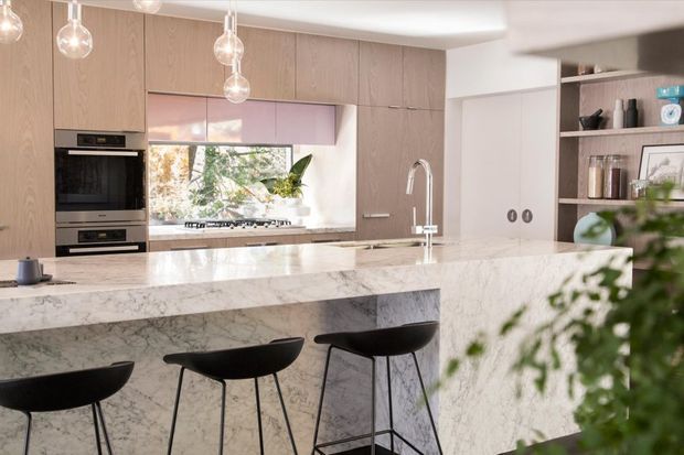 Bright contemporary kitchen featuring a large marble island with waterfall edges and integrated seating for four, pale timber cabinetry, built-in stainless-steel appliances, a white splashback, and a sculptural chrome tap. Pendant globe lights hang above the island, while open shelving displays kitchenware and plants.