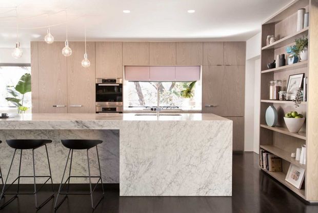 A contemporary kitchen with a large marble-look island and two black bar stools, full-height light timber cabinetry, integrated appliances, a sink beneath a window, pendant lights, dark timber flooring, and open display shelving along the right wall. The space is bright and refined, with greenery visible through the glazing.