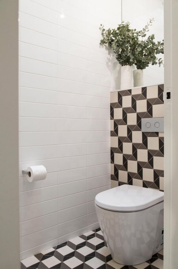 A compact modern bathroom or powder room featuring a wall-hung toilet, white horizontal wall tiles, and a bold black, white, and grey geometric tiled floor and concealed cistern feature wall. A chrome flush plate and toilet-roll holder provide simple fittings, while two tall white vases with leafy branches add a natural decorative accent.