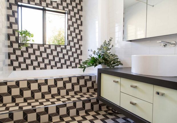 A bright contemporary bathroom featuring a geometric black, white and grey patterned tile wall and floor, a built-in bathtub, and a floating vanity with a dark benchtop, pale cabinetry and a white vessel basin. A black-framed window provides natural light, while a wall-mounted mirrored cabinet and small indoor plants complete the space.