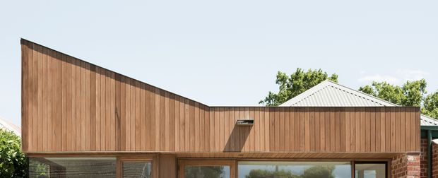 Exterior view of a contemporary residential building with a long, angled timber-clad upper facade and broad glazed openings below. A corrugated metal gabled roof and partial brickwork are visible behind the warm vertical timber screen, set against a clear sky and greenery.