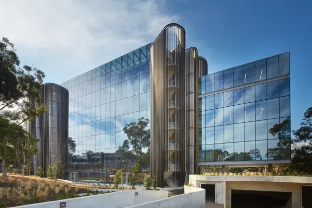 Exterior view of a contemporary multi-storey commercial building with a reflective glass curtain wall and distinctive rounded vertical circulation towers clad in dark metal fins. Landscaped grounds, concrete retaining walls, and an integrated lower-level vehicle entry are visible in the foreground.