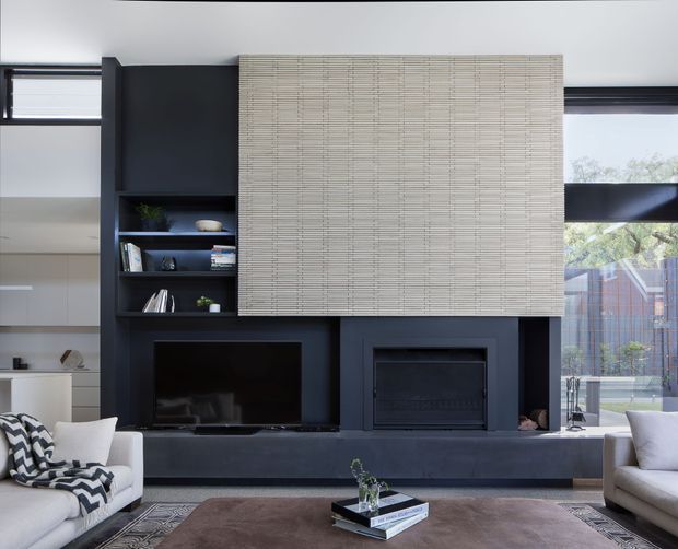 Contemporary living room centred on a dramatic dark built-in media and fireplace wall. Open shelving displays a few decorative objects, while a large woven light-toned feature panel conceals the upper portion of the wall. Sofas, a coffee table and rug occupy the foreground, with extensive glazing and views to greenery visible on the right.