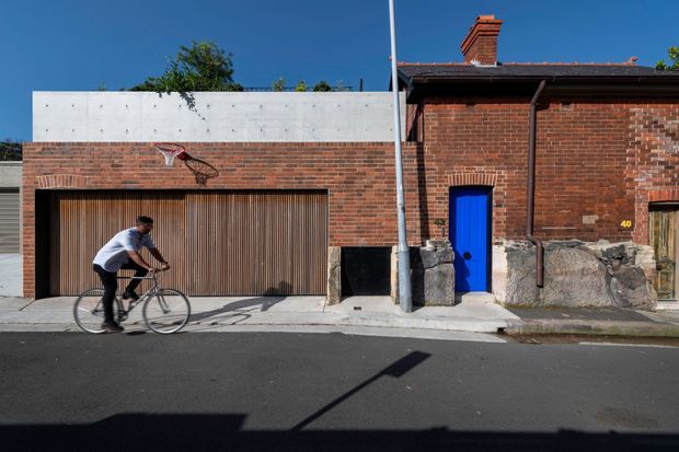 Street-facing view of a compact brick building or residence with a contemporary white rendered upper section, a large vertically slatted timber garage door, a vivid blue pedestrian door, exposed masonry and a traditional brick chimney. A cyclist passes in the foreground on a paved urban street.