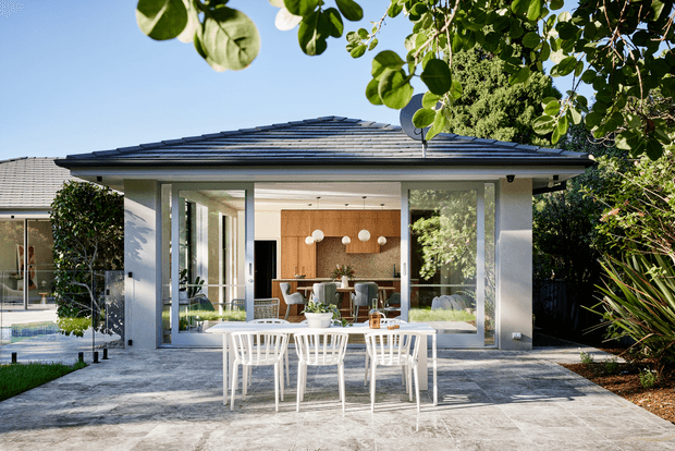 Bright exterior view of a modern single-storey pavilion-style home opening onto a broad paved patio. Floor-to-ceiling glazing reveals an open-plan dining and living area with warm timber cabinetry, while a white outdoor dining table and chairs occupy the foreground. The low hipped roof, pale rendered walls, extensive glass, landscaped garden, and swimming pool edge create a clean indoor-outdoor setting.