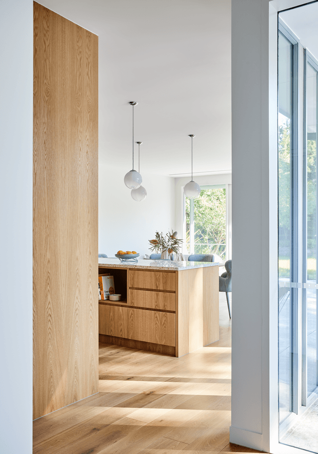 Bright, minimalist open-plan kitchen and dining area viewed through a doorway. The space features extensive light oak wall panelling and flooring, a central timber kitchen island with integrated drawers and open shelving, pale stone benchtops, white walls and ceiling, sculptural globe pendant lights, and large glazed doors or windows bringing in abundant natural light.