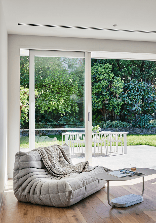 Bright contemporary living room with a low, plush grey modular sofa and knit throw positioned beside full-height sliding glass doors. The room has warm timber flooring, a slim dark coffee table, white walls and ceiling, and views to a leafy garden with an outdoor dining setting beyond the glazing.