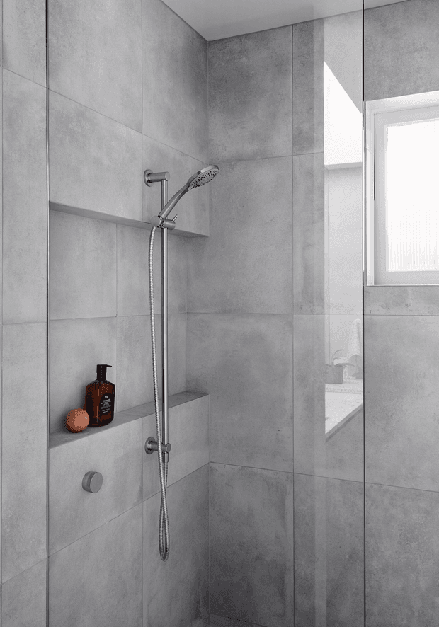 A compact modern walk-in shower enclosed by clear glass, with large-format grey concrete-look wall tiles, a chrome handheld shower rail, recessed tiled niches, and a small frosted window. A mirrored or reflective panel reveals part of the adjacent vanity area. The restrained monochrome palette is accented by a dark soap bottle and an orange decorative object.