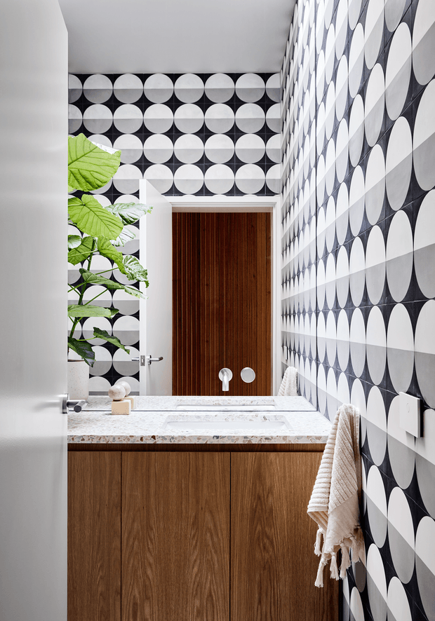 A compact, high-impact bathroom viewed through an open door. The room features bold black-and-white circular patterned wall tiles, a white terrazzo-style vanity benchtop with integrated basin, warm vertical timber vanity cabinetry, a matching timber door with round hardware, and a leafy plant visible near the rear. A textured neutral towel hangs on the right wall. The design combines graphic patterning with warm natural wood and crisp white surfaces.