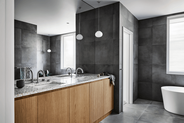 A contemporary bathroom vanity area with a long light-wood fixed cabinet, speckled stone benchtop, twin basins and chrome tapware. Dark large-format wall and floor tiles create a dramatic backdrop, with a freestanding white bathtub visible to the right. A large mirror reflects the vanity and pendant lights, while windows provide bright natural light.