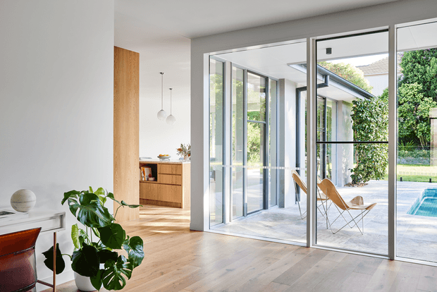 Bright contemporary open-plan interior with pale timber flooring, white walls, a timber kitchen and large floor-to-ceiling glazed doors opening toward a patio and swimming pool. A leafy indoor plant and minimal furniture sit in the foreground, while sculptural pendant lights and a warm timber joinery wall add detail.