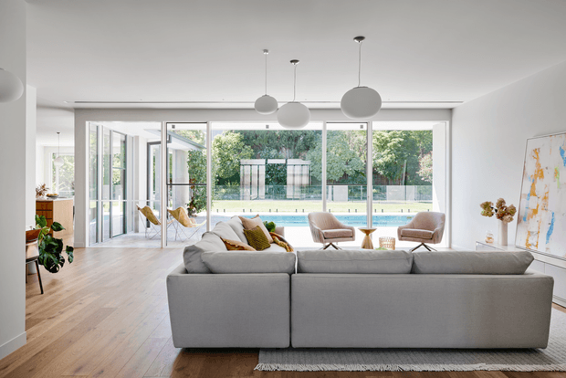 Bright, spacious contemporary living room with a large grey sectional sofa facing floor-to-ceiling glazed doors and windows. The open-plan space has light timber flooring, white walls and ceiling, sculptural globe pendant lights, occasional lounge chairs, indoor plants and abstract artwork. Through the glazing, a swimming pool and landscaped garden are visible.