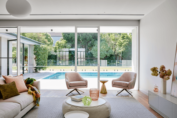 Bright contemporary living room with floor-to-ceiling glazing overlooking a swimming pool and landscaped garden. The room features a pale sectional sofa, two upholstered lounge chairs, a round upholstered coffee table, a low white media console, light timber flooring, a large neutral rug, and a sculptural ceiling light. The space has a clean, airy indoor-outdoor connection and abundant natural light.