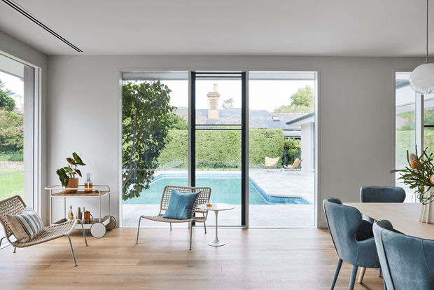 Bright contemporary living and dining space with pale timber flooring, white walls, full-height glazing and slim dark window frames overlooking a swimming pool and landscaped garden. A woven lounge chair, blue upholstered armchairs, compact side tables, a small drinks trolley and indoor plants create a refined, airy interior.