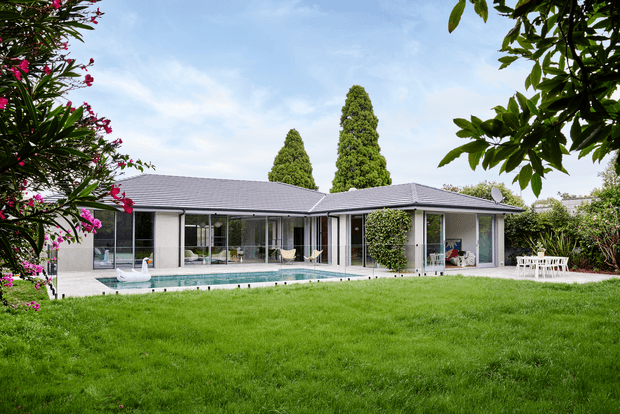 Single-storey contemporary residence with a low-pitched hipped roof, pale rendered walls, extensive floor-to-ceiling glazing, and a covered outdoor living area arranged around a rectangular swimming pool. The home opens onto a broad manicured lawn and landscaped garden, with mature trees and flowering shrubs framing the view.