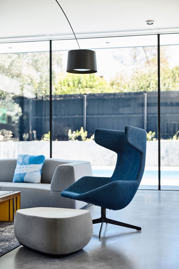 Bright contemporary living room with a sculptural navy-blue lounge chair and matching ottoman, a low grey sofa, patterned cushion, and a small timber side table. Full-height black-framed glazing opens the room visually to a courtyard or pool area, while a simple black pendant light hangs from the ceiling. The space has a polished, uncluttered, indoor-outdoor character.