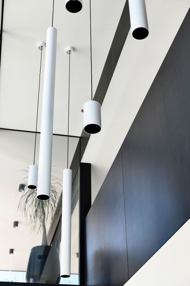 Close-up upward view of a contemporary interior featuring multiple white cylindrical pendant lights suspended from a white ceiling. Strong black geometric ceiling and wall elements create bold diagonal lines, while a tall potted plant and dark reflective wall panel are visible below. The composition emphasizes minimalist lighting, high contrast, and a dramatic double-height feel.
