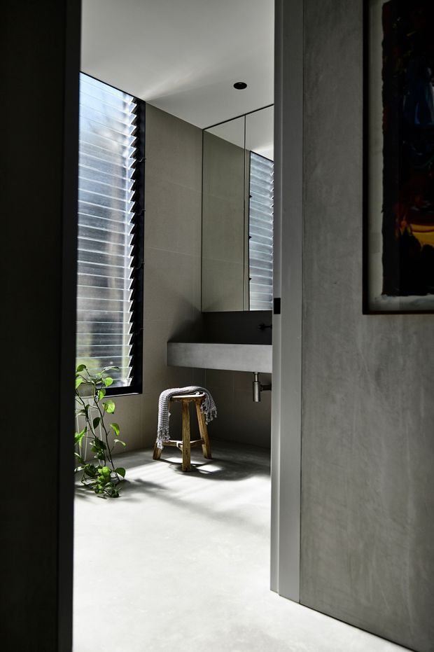 A minimalist contemporary bathroom viewed through a doorway, featuring a floating rectangular concrete or stone basin with a wall-mounted tap, mirrored cabinetry, grey microcement-like walls and floor, and a tall louvred window providing strong natural light. A small wooden stool with a draped textile and a potted plant add warmth to the otherwise restrained space.