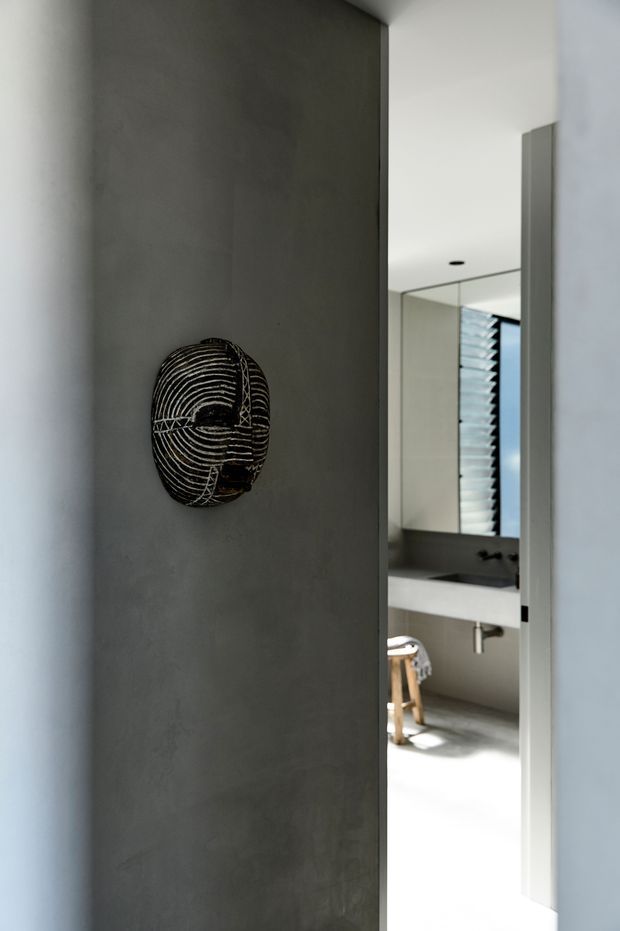 A minimalist interior view looking through a narrow opening into a contemporary bathroom. The foreground features a dark grey textured feature wall with a sculptural black-and-white wall mask. In the bathroom beyond, a large mirror, floating dark vanity, white basin, wall-mounted tapware, and a timber stool are visible. The palette is monochromatic with strong natural light and clean architectural lines.