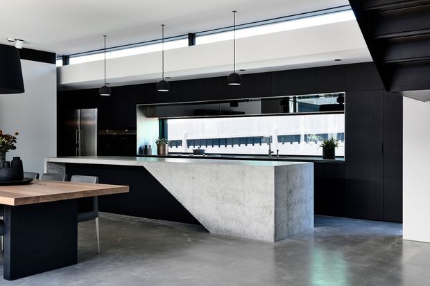 Contemporary open-plan kitchen and dining area with a dramatic black built-in kitchen wall, large concrete island with waterfall-style end, polished grey concrete floor, timber dining table, dark cabinetry, pendant lights, and a long horizontal glazed opening providing reflected views and daylight.
