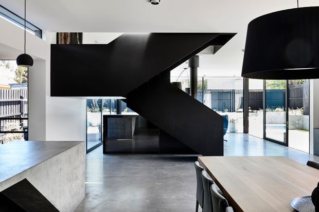 Open-plan contemporary interior dominated by a sculptural black enclosed staircase with angular geometry. The space combines polished concrete flooring, white walls and ceilings, expansive black-framed glazing, a concrete kitchen island, and a timber dining table. A pool and landscaped courtyard are visible beyond the glazing.