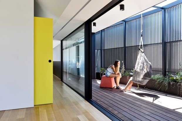 Bright contemporary indoor-outdoor living area with a timber-floored corridor opening onto a covered deck. Full-height black-framed glazing separates the interior from the deck, which features a hanging hammock chair, colourful cube seating, and built-in planters behind a vertical slatted privacy screen. A vivid yellow pivot door and pale timber flooring add a bold accent to the minimalist space.