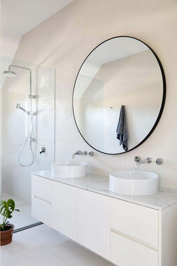 Bright contemporary bathroom with a double floating vanity, two white vessel basins, pale marble-look benchtop, wall-mounted chrome tapware, large rounded black-framed mirror, and a glass-enclosed shower. Light neutral wall and floor finishes create a clean, airy appearance, with a small potted plant adding greenery.