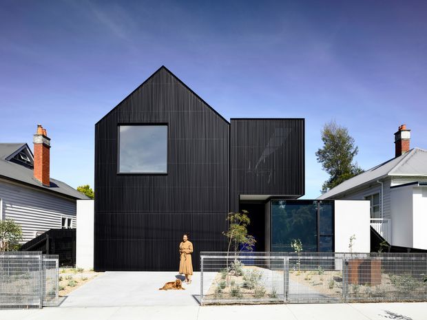 Front exterior view of a striking contemporary two-storey residence with a steep gabled black vertically ribbed facade and projecting upper volume. Large dark-framed glazing sits beneath the cantilevered section, while low pale walls, metal fencing, sparse landscaping, and a concrete driveway define the frontage. Traditional neighbouring houses and chimneys contrast with the new dark modern building.