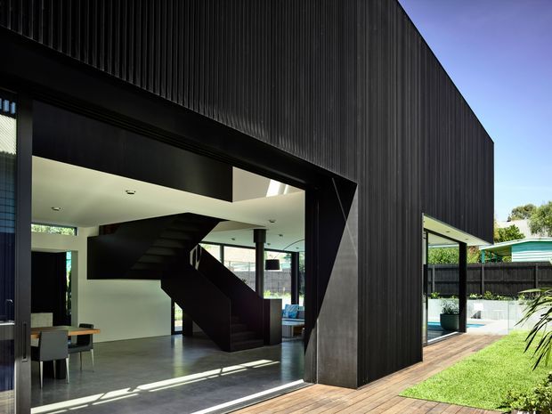 Contemporary two-storey residence with a dramatic black vertically ribbed exterior and a large open glazed opening. The view looks from the timber deck into a bright open-plan interior featuring a sculptural black staircase, white walls, polished concrete flooring, and glimpses of dining and living areas. A pool courtyard and landscaped garden are visible to the side.