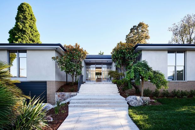Front exterior view of a low-slung, single-storey contemporary residence with two symmetrical wings framing a recessed glazed entry. The facade combines smooth light plaster, dark horizontal lower cladding, and warm brickwork, with broad flat rooflines and large windows. A wide pale paved pathway and steps lead through a carefully landscaped garden with clipped shrubs, palms, rocks, and lawn.
