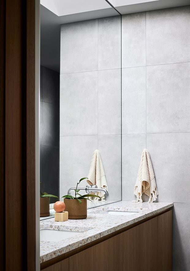 A contemporary bathroom vanity viewed through a timber-framed doorway. Large-format light grey wall tiles form a clean backdrop, with a mirrored wall reflecting the vanity, folded towels and plants. The double vanity has a pale terrazzo-style stone benchtop, integrated white basins, brushed metal tapware and dark timber-look cabinetry. A skylight or ceiling opening provides soft natural light.