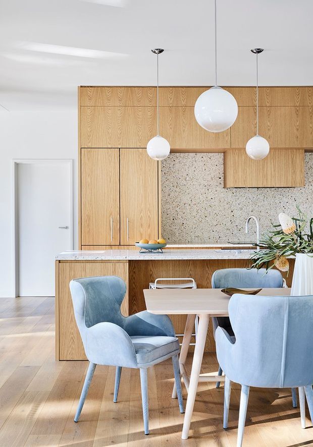Bright open-plan kitchen and dining area with a full-height light timber kitchen wall, integrated cabinetry, speckled stone splashback, island bench, and pale timber flooring. A light wood dining table is surrounded by soft blue upholstered chairs, while three white spherical pendant lights hang above the kitchen and dining zones. The space has clean contemporary detailing and abundant natural light.
