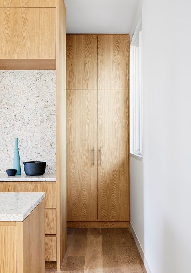A narrow, bright kitchen passage featuring extensive light timber cabinetry, a tall built-in storage cupboard, pale terrazzo-style splashback, and pale stone benchtops. A timber floor runs through the space, with a white wall and window along the right side. Minimal dark vessels provide small decorative accents.