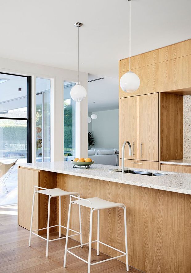 Bright contemporary kitchen with a large light-oak island and full-height cabinetry, pale stone benchtops, integrated sink and curved chrome tap, two white bar stools, and spherical pendant lights. Full-height glazing opens toward an outdoor area, while a living room is visible beyond the kitchen.