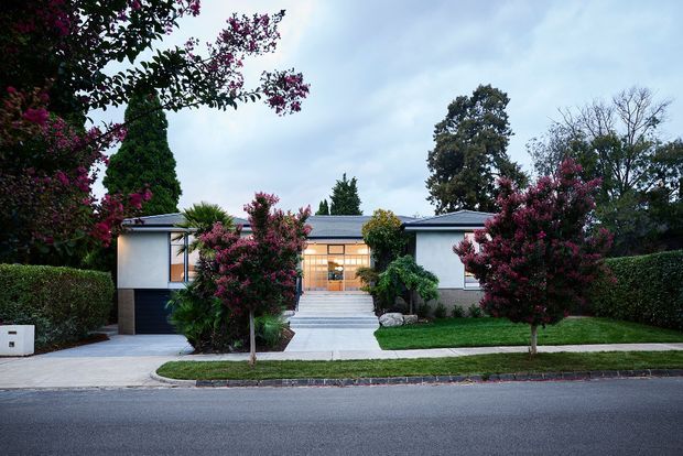 Front exterior view of a low, single-storey modernist house with a broad low-pitched roof, symmetrical composition, pale rendered walls, dark timber or metal garage door, large glazed entry, and a straight concrete path leading from the street. Mature hedges and flowering trees frame the manicured front garden.
