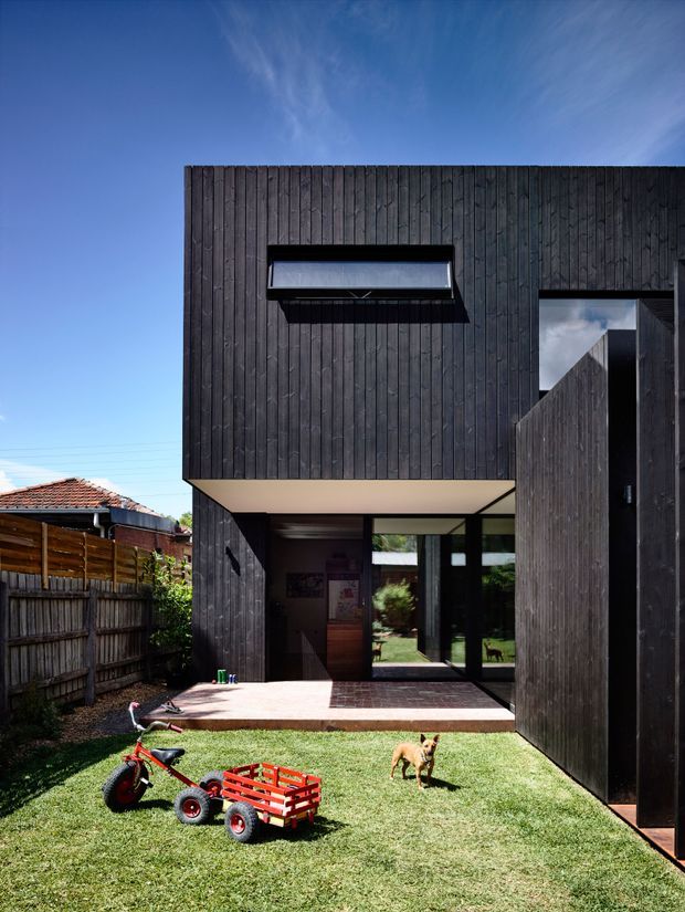 Exterior view of a contemporary two-storey house with a dark vertically timber-clad facade, a projecting upper volume, and a recessed ground-floor entry opening onto a small lawn and paved terrace. Large glazed doors connect the interior to the garden. A tall timber privacy gate or screen frames the right side, while neighbouring low-rise housing and fencing are visible to the left. The image is captured in bright daylight with a strong blue sky.