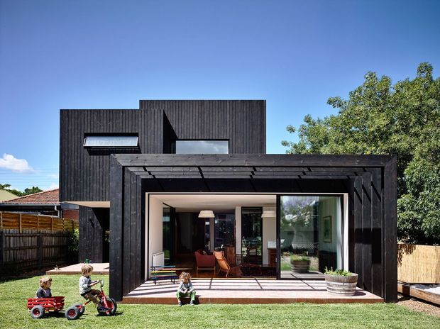 Exterior view of a contemporary two-storey suburban house with a dark vertically clad upper volume and a projecting framed outdoor living area. Large sliding glass doors open the ground-floor living space onto a paved terrace, with children’s toys and garden elements in the foreground. The facade has strong rectilinear geometry, deep timber framing, and a flat roofline.