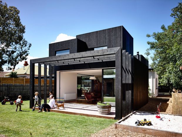 Exterior view of a contemporary multi-level residential extension with dark timber cladding, strong rectilinear volumes, and a covered outdoor terrace. Large glazed openings reveal a bright living area inside, while the landscaped backyard includes lawn, children's play space, and a sandpit.