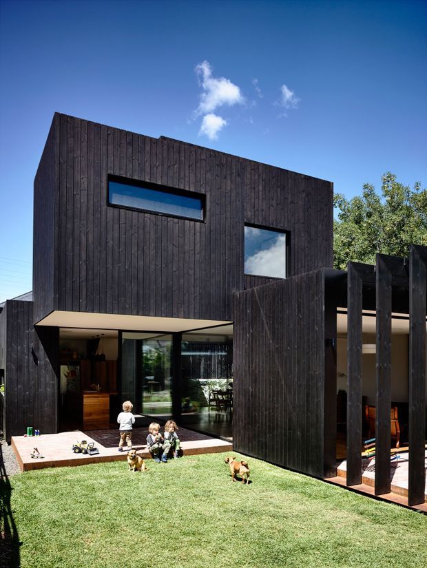 Exterior view of a contemporary two-storey house with a bold dark vertical timber-clad facade, irregular projecting volumes, narrow horizontal and square windows, and a sheltered ground-level living area opening onto a paved lawn. A dark timber screen with tall vertical fins creates privacy and defines an outdoor terrace. Children and dogs are present in the garden.
