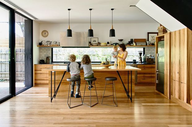 A bright, open-plan contemporary kitchen with a large timber island and three stools, extensive warm wood cabinetry, white tiled backsplash, black-framed glazing, and three black pendant lights. Two children sit at the island while an adult stands behind it. The room has a light timber floor and opens toward a glazed outdoor area.