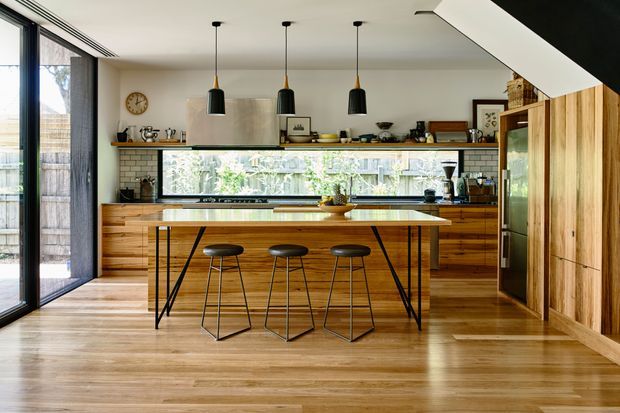 A warm, contemporary kitchen with extensive timber cabinetry, a long central island with seating for three, open shelving, a tiled backsplash, black pendant lights, and wide glazed doors along the left side. The room has timber flooring and a bright, connected indoor-outdoor feel.