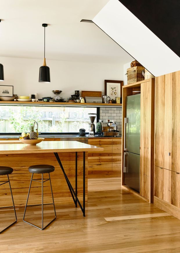Bright, compact contemporary kitchen with extensive warm timber cabinetry and matching timber flooring. A central island with a pale stone benchtop and slim black metal supports provides seating for two on dark stools. The rear wall includes a long horizontal window, open display shelving, coffee-making appliances, and a white subway-tile backsplash. Full-height timber pantry cabinetry and an integrated refrigerator occupy the right side, while black pendant lights and open shelving add contrast.